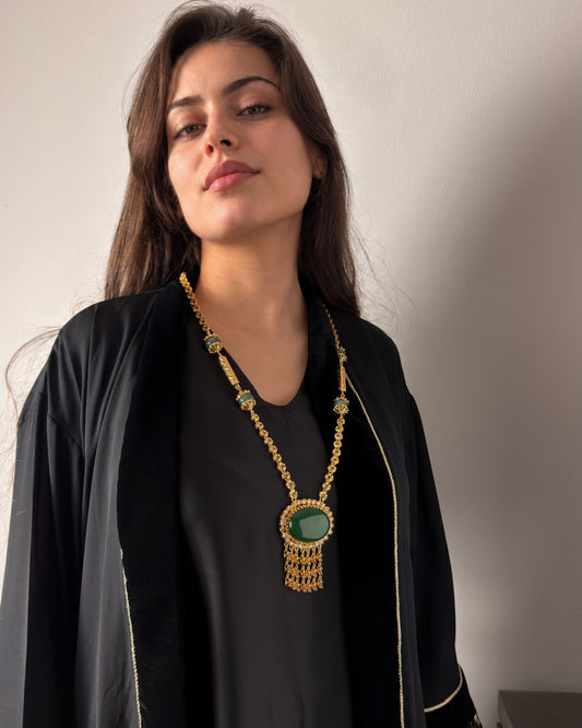 Portrait d’une femme en tenue noire portant un long sautoir doré à cabochon ovale vert et pampilles. Le bijou est visible devant un mur beige, en lumière naturelle.