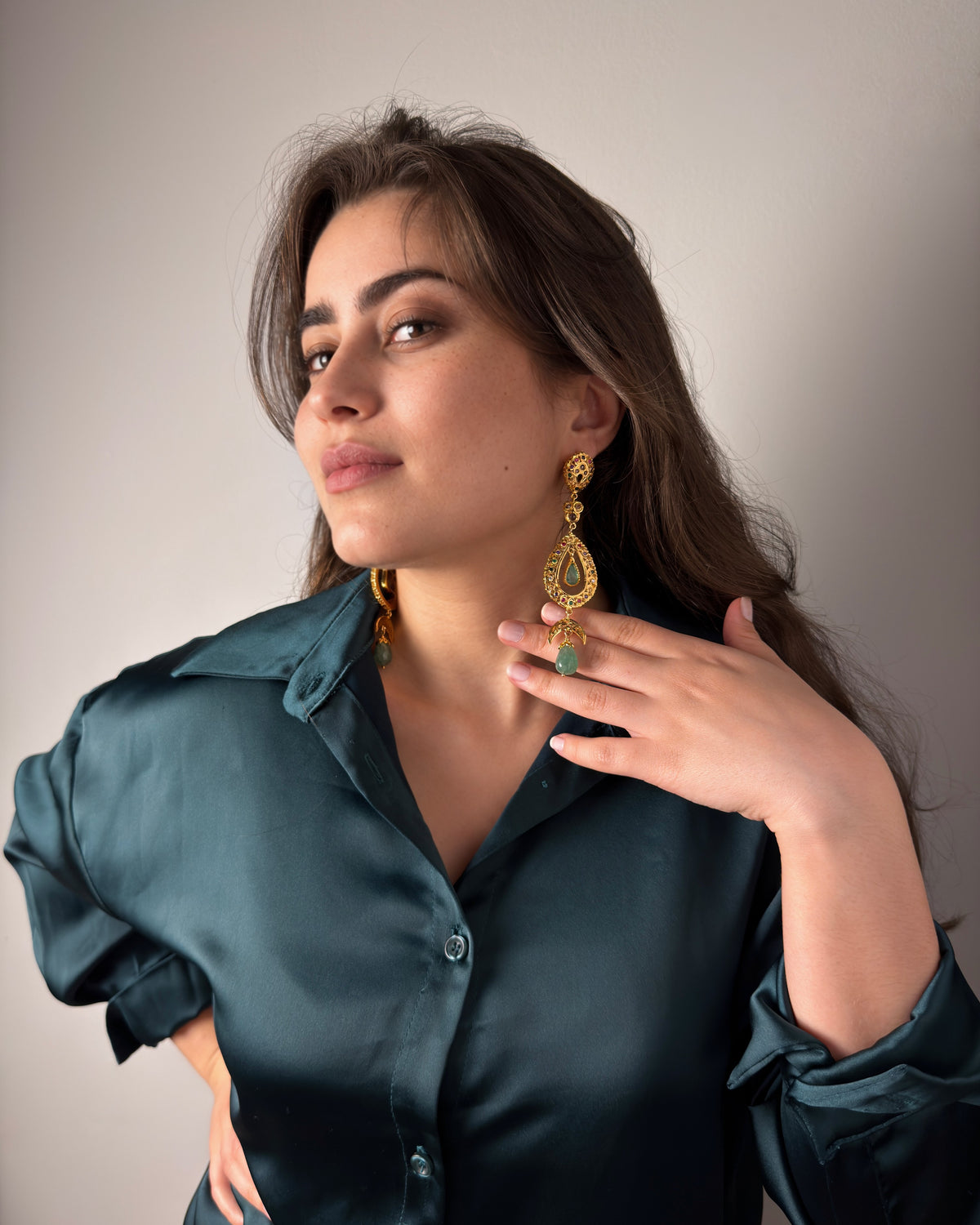 Model wearing long Moroccan drop earrings with green cabochons and a teal satin shirt, with her hand beside one earring.