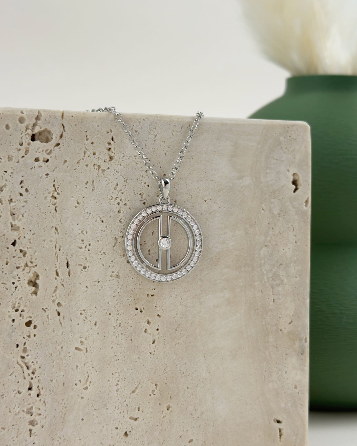 Silver moon pendant necklace displayed on beige travertine stone with a green vase in the background for a refined Amuris jewelry look.