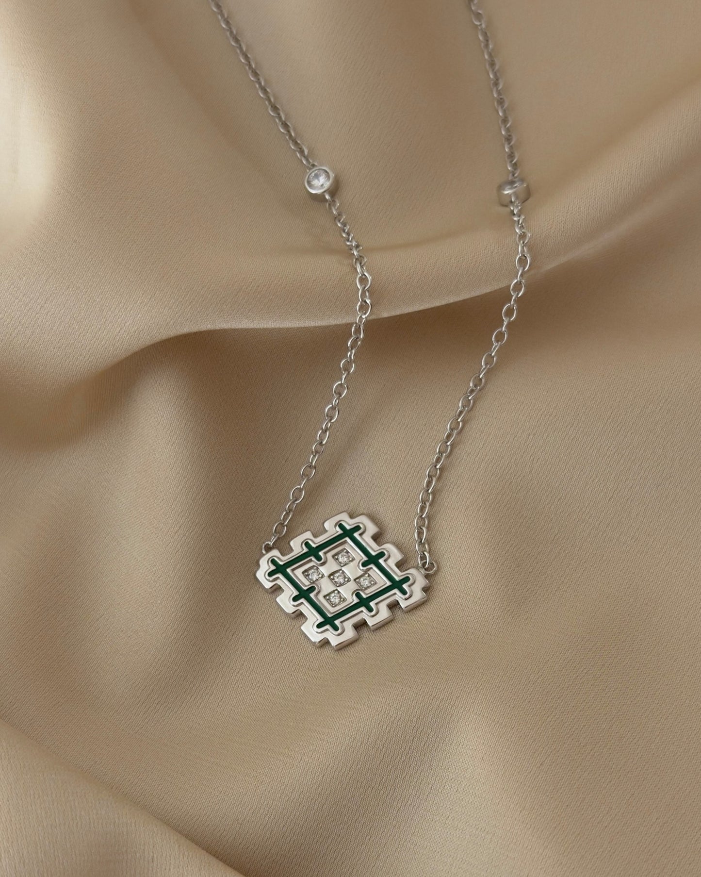 Close-up of a rhodium-plated silver Lion’s Paw necklace with green enamel and sparkling cubic zirconia details, styled on soft beige fabric.