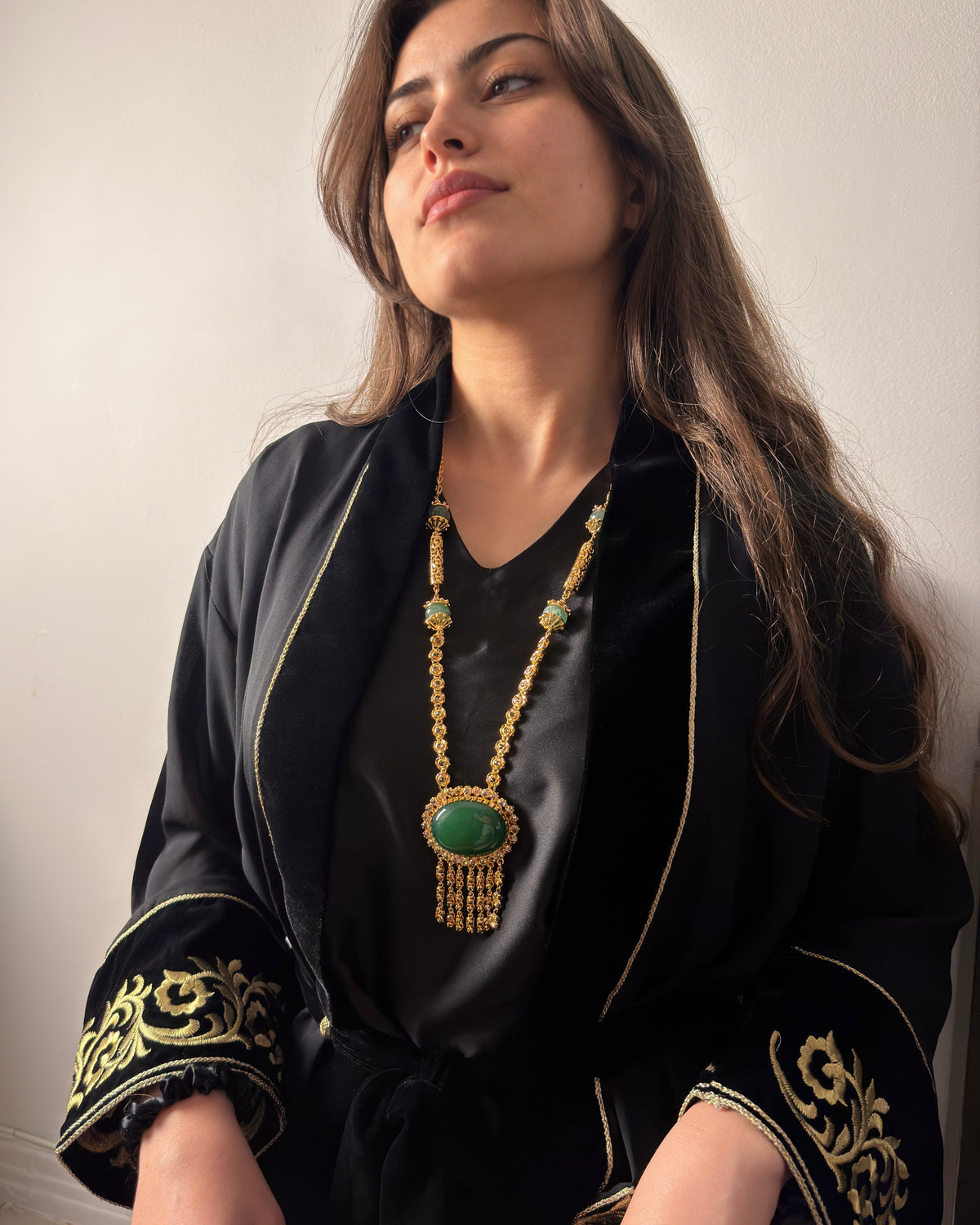 Femme assise, le regard tourné sur le côté, portant un long sautoir doré à cabochon vert et pampilles sur une tenue noire aux manches brodées de doré.