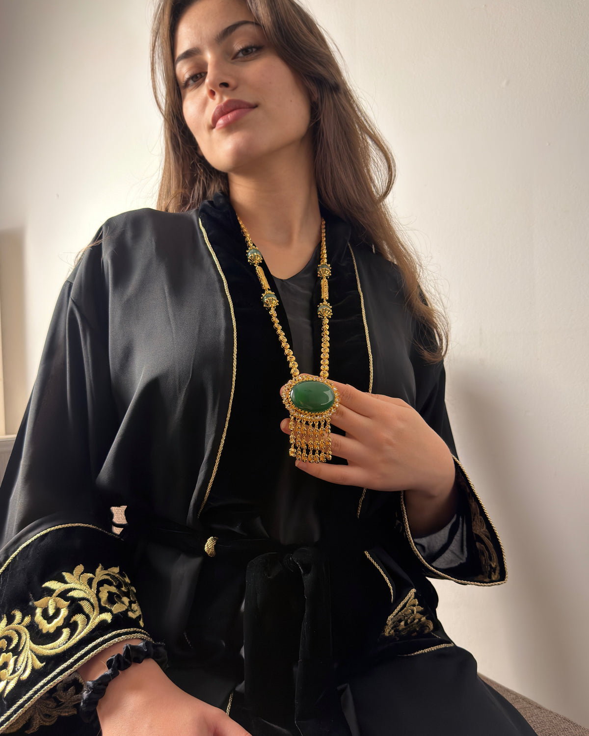 Femme assise en tenue noire brodée de motifs dorés, tenant délicatement le pendentif vert de son sautoir. Des pampilles dorées retombent devant ses doigts.