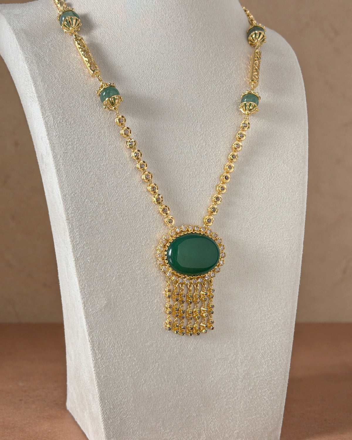 Vue légèrement oblique d’un sautoir doré sur un présentoir beige, montrant le cabochon vert, les rosaces et les pampilles sur un fond terracotta.