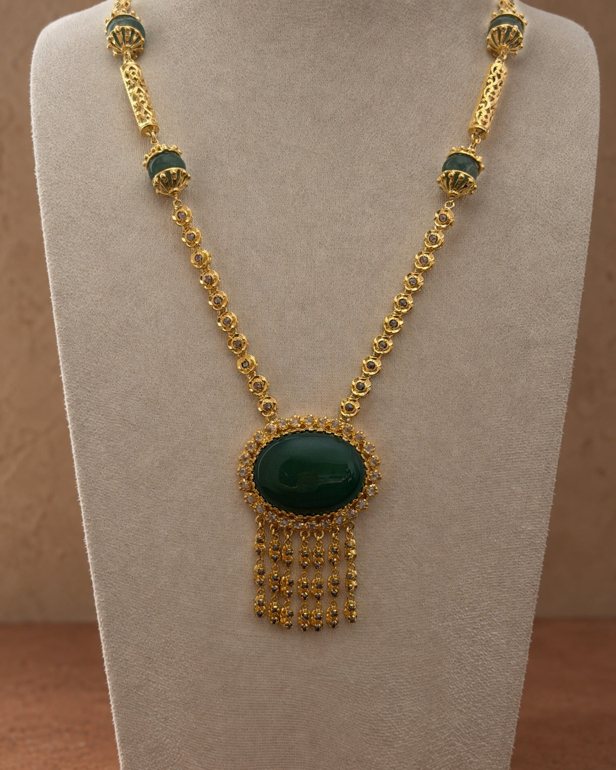 Sautoir doré sur un présentoir beige, avec cabochon ovale vert, rosaces scintillantes et pampilles. Des éléments verts ponctuent la chaîne, sur un fond terracotta.